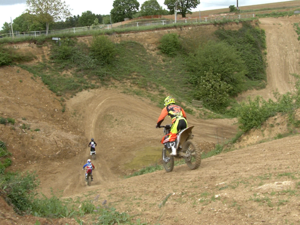 Trois motards en tenue de motocross descendent une piste de terre en pleine nature, entourée de collines verdoyantes.