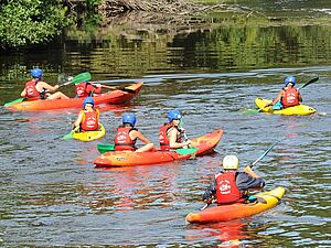 Un groupe de personnes en gilets rouges et casques colorés fait du kayak sur une rivière calme entourée de verdure.