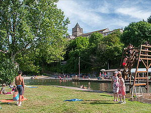 Des personnes profitent d’un après-midi ensoleillé au bord d’un plan d’eau, entre baignade, détente sur l’herbe et jeux près d’un plongeoir en bois, avec une église en arrière-plan dominant la scène.