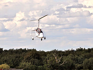 Un autogire blanc avec deux personnes à bord vole au-dessus d’une forêt sous un ciel partiellement nuageux.